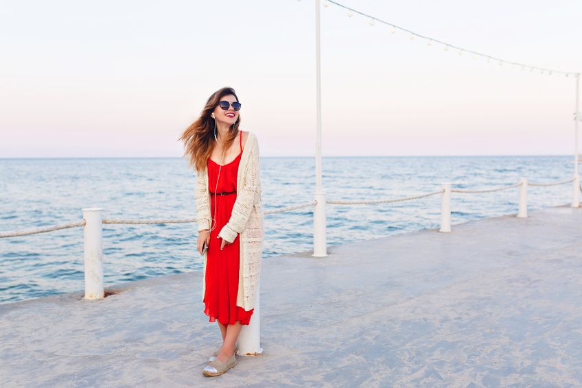 Beautiful girl in red dress and white jacket stands on a pier, smiles, and listens to music on earphones on a smartphone. Girl has ombre hair. She wears light brown espadrilles and dark sunglasses.